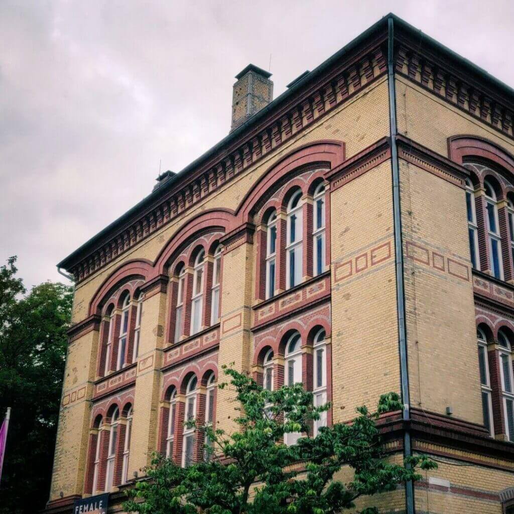 Historisches Gebäude mit roten Ziegeln und großen Fenstern, umgeben von Bäumen, unter einem bewölkten Himmel.
