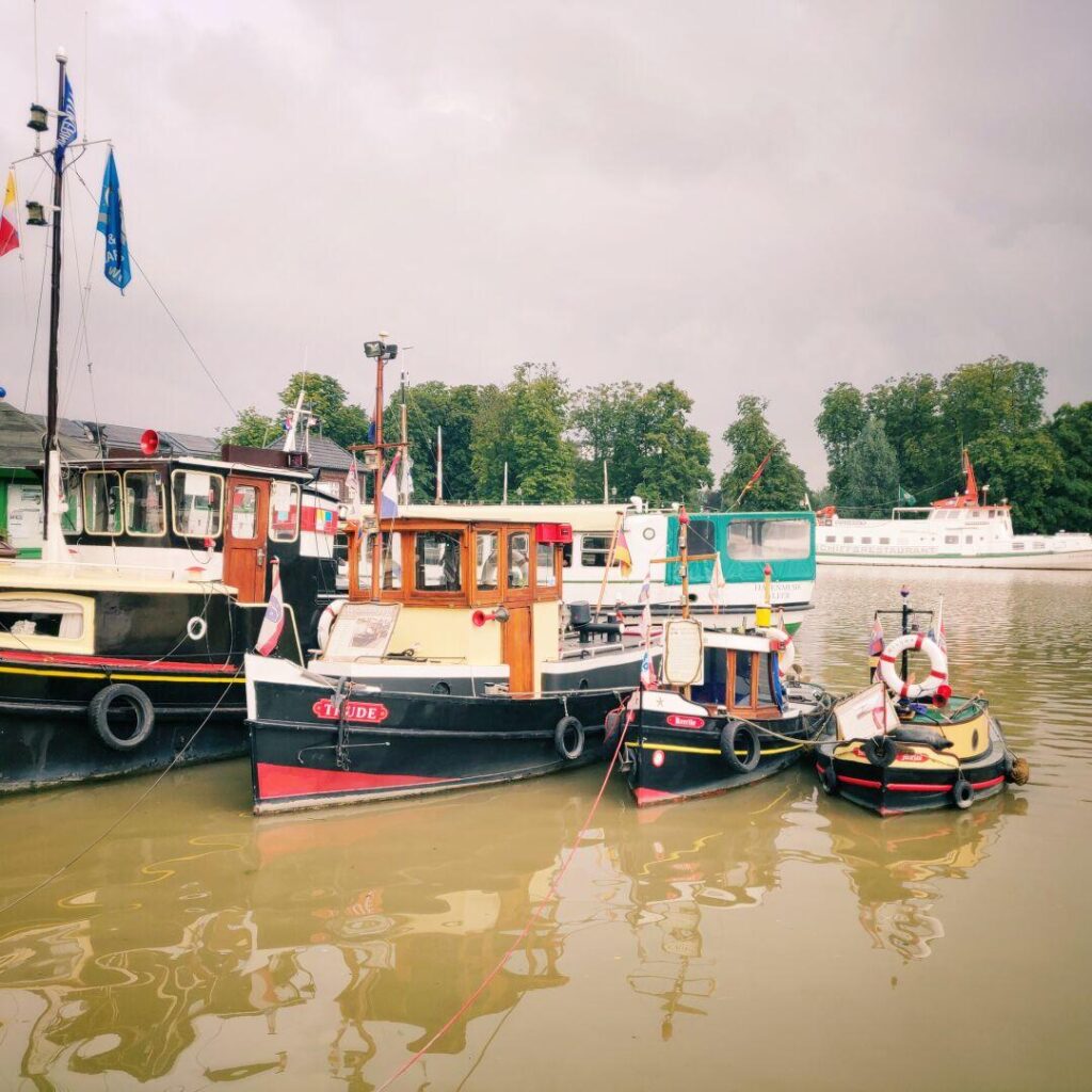 Mehrere bunt dekorierte Boote liegen an einem ruhigen Hafen, umgeben von grünen Bäumen und einem bewölkten Himmel, während im Hintergrund ein größeres Schiff vorbeifährt.