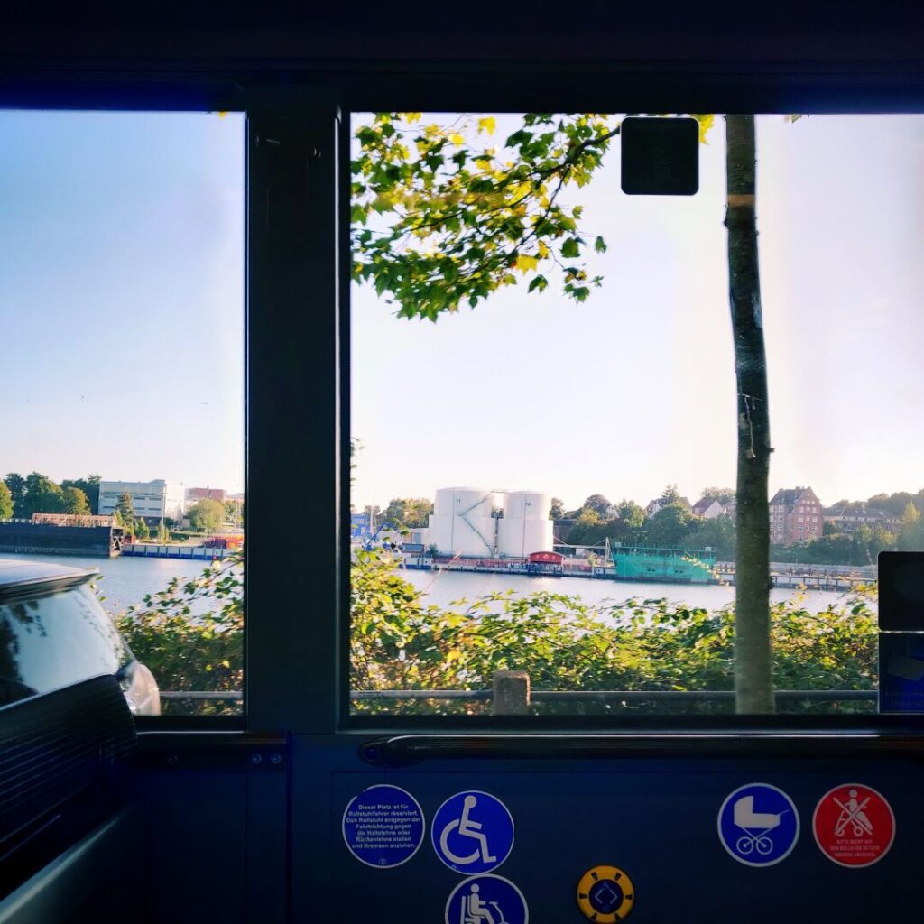 Blick aus einem Busfenster auf eine malerische Uferlandschaft mit einem Fluss, umgeben von Bäumen, Gebäuden und zwei großen Lagertanks im Hintergrund, während die Sonne am blauen Himmel scheint.
