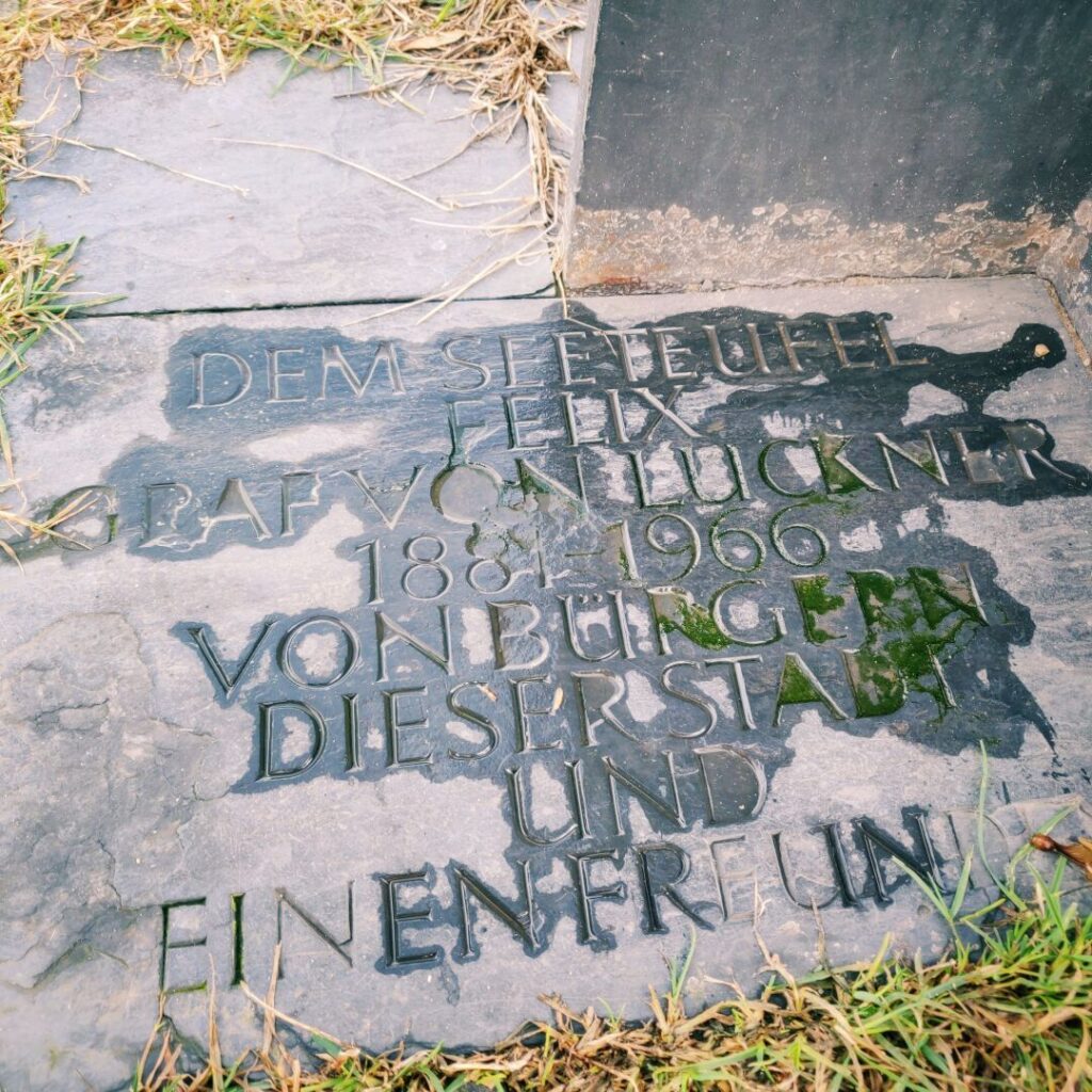Grabstein mit der Inschrift zu Ehren von Felix Graf von Luckner (1884-1966), der in der Stadt von Burg und seiner Freundin gedenkt, umgeben von Gras und Erde.