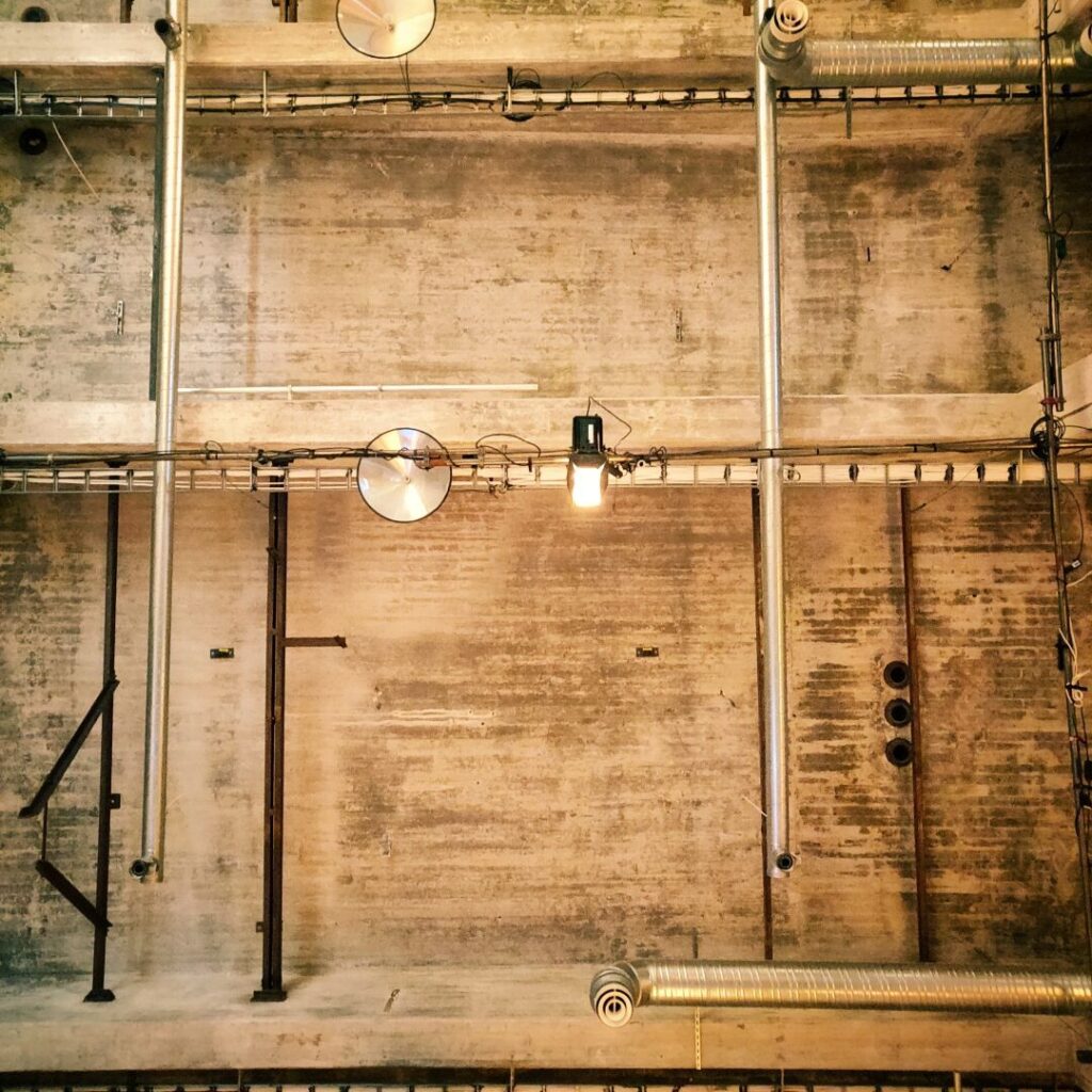 Die Aufnahme zeigt eine Decke mit sichtbaren Rohren, Kabeln und Lampen in einem industriellen Raum, wobei die Wände aus grobem Beton bestehen und eine minimalistische, moderne Atmosphäre vermitteln.