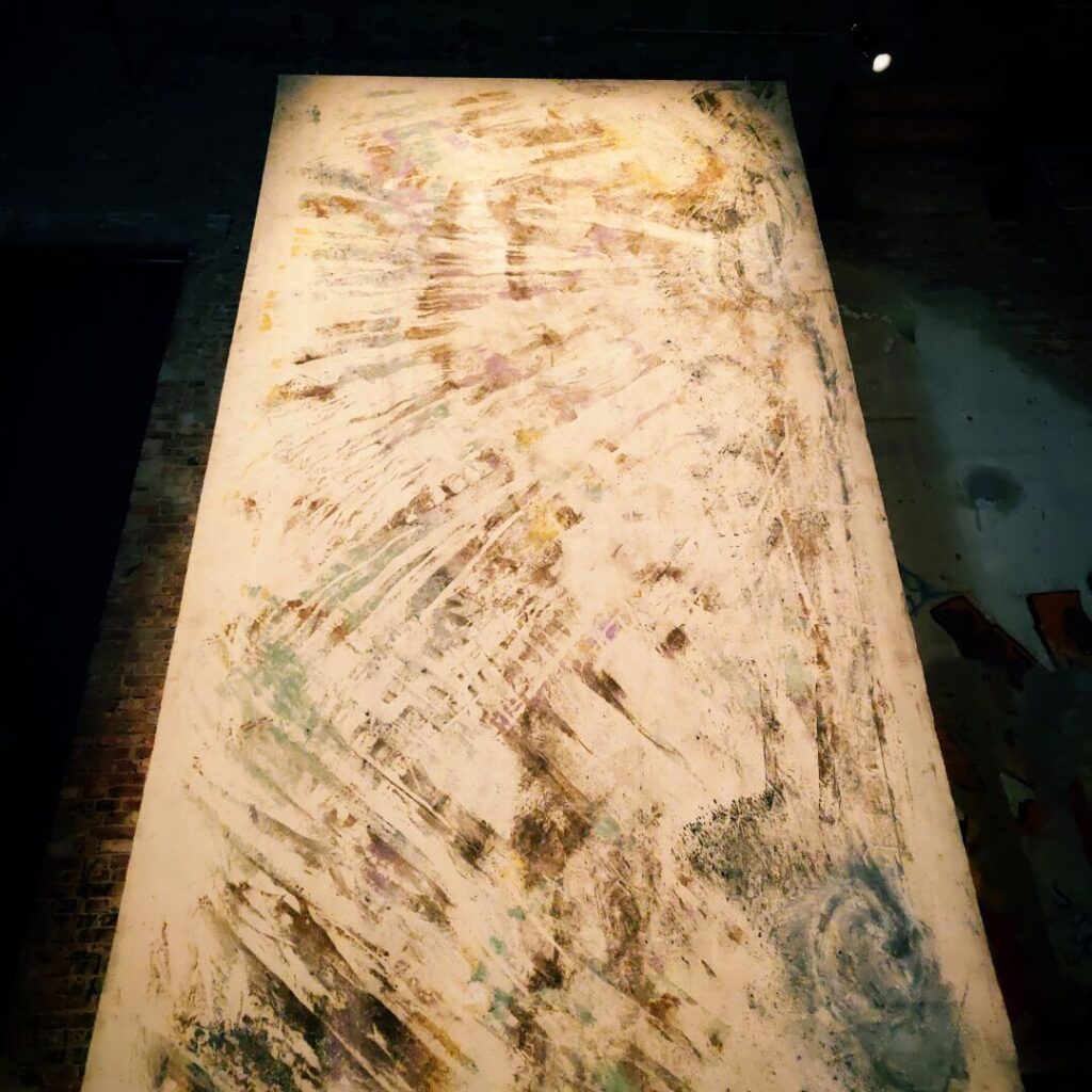 Eine große, abstrakte Leinwand mit sanften Farbtönen und dynamischen Pinselstrichen, die in einem dunklen Raum beleuchtet wird, vermittelt eine Atmosphäre von Kreativität und Kunst.