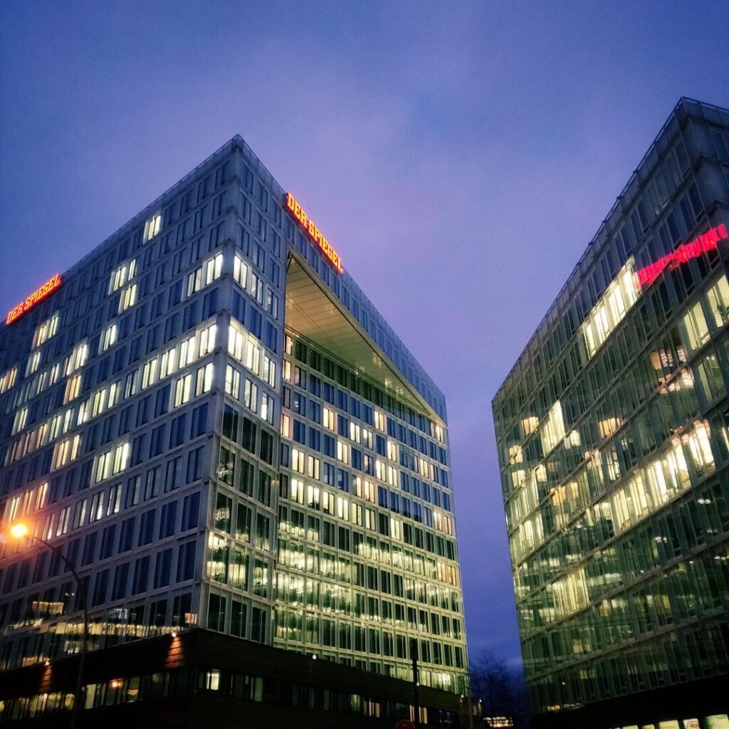 Moderne Bürogebäude mit Glasfassade und leuchtenden Fenstern, die den Schriftzug "DER SPIEGEL" tragen, vor einem bewölkten Abendhimmel.