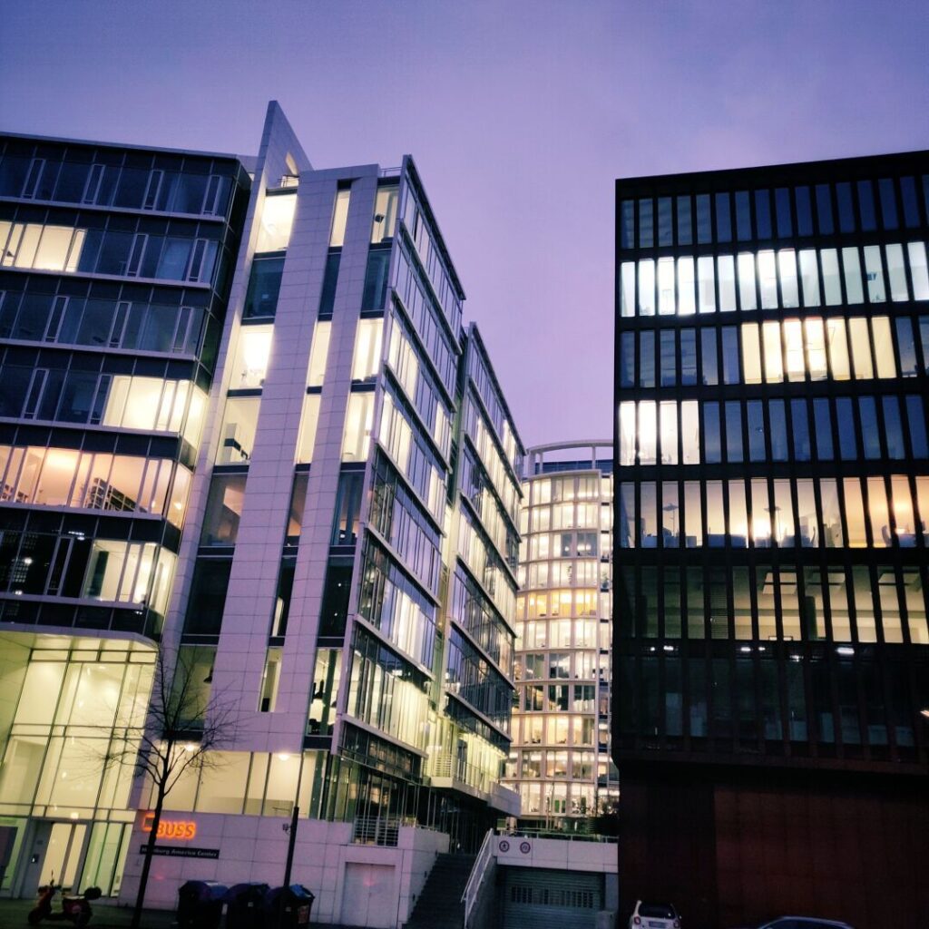 Moderne Bürogebäude mit großen Glasfassaden und beleuchteten Büros, aufgenommen bei Dämmerung, zeigen eine städtische Architektur in einem urbanen Umfeld.