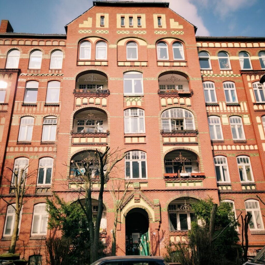Historisches Backsteingebäude mit mehreren Balkonen, großen Fenstern und dekorativen Elementen, umgeben von Bäumen und Sträuchern, aufgenommen an einem sonnigen Tag.