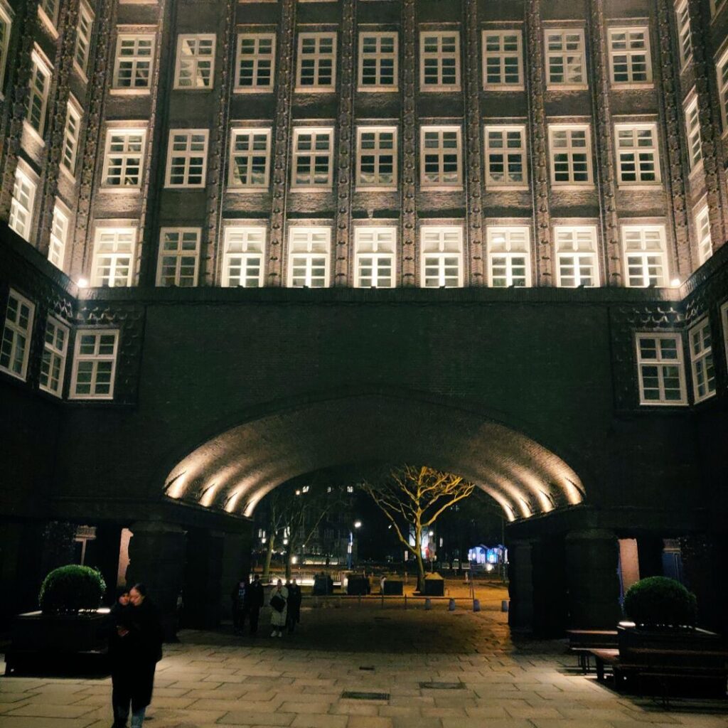 Eine nächtliche Aufnahme eines historischen Gebäudes mit einem großen, beleuchteten Bogen, umgeben von Fenstern und einem gepflasterten Platz, auf dem Menschen spazieren gehen und Bäume im Hintergrund sichtbar sind.