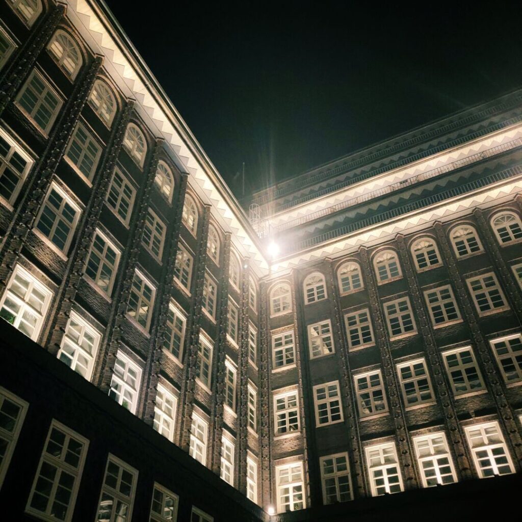 Die Aufnahme zeigt eine beeindruckende historische Backsteinarchitektur bei Nacht, beleuchtet von warmem Licht, das die Fenster und Details der Fassade hervorhebt und eine stimmungsvolle Atmosphäre schafft.