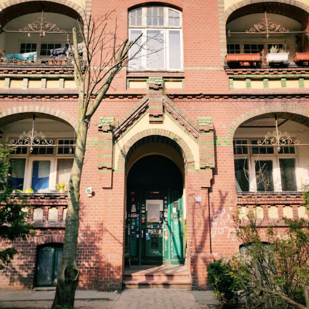 Fassade eines historischen Backsteingebäudes mit grünen Türen, verzierten Fenstern und einem kleinen Baum im Vordergrund, das eine einladende Atmosphäre ausstrahlt.