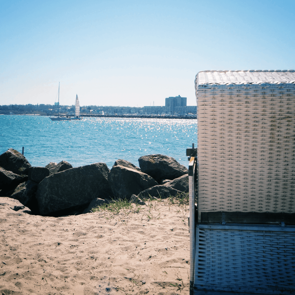 Ein Strandkorb steht am Ufer, während sanfte Wellen auf die Steine treffen und im Hintergrund ein Segelboot sowie die Silhouette einer Stadt unter blauem Himmel sichtbar sind.