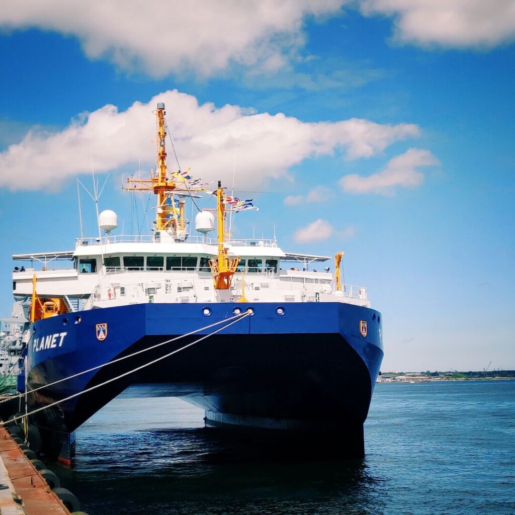 Ein großes, blaues Forschungsschiff mit dem Namen "PLANET" liegt am Hafen, umgeben von klarem Wasser und einem strahlend blauen Himmel mit einigen Wolken.