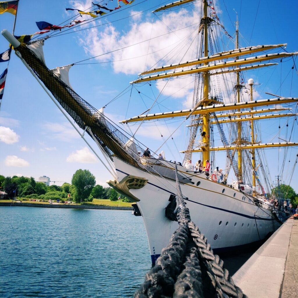 Ein prächtiges Segelschiff mit gelben Masten liegt am Ufer eines ruhigen Gewässers, umgeben von grünen Bäumen und einem blauen Himmel mit wenigen Wolken, während Menschen an Bord und am Steg zu sehen sind.
