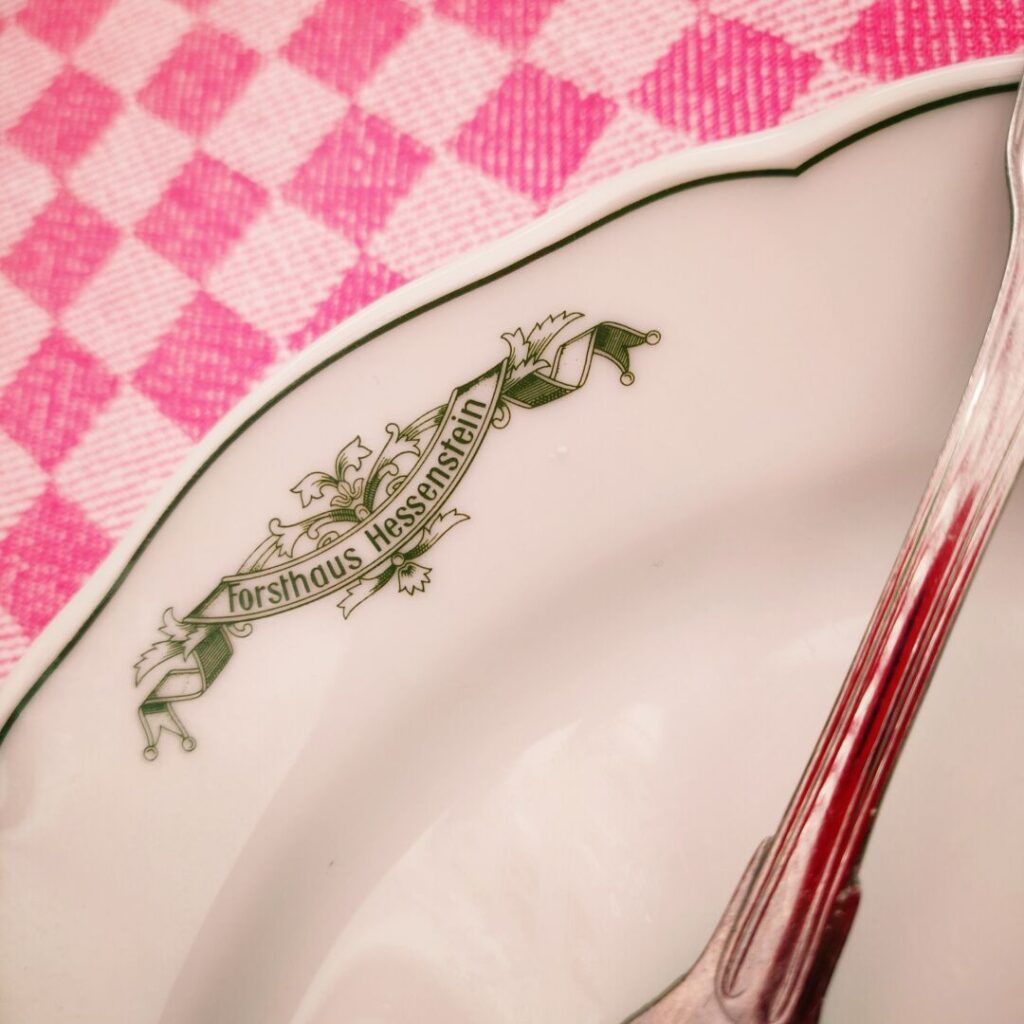 Ein elegantes, weißes Porzellanteller mit dem Schriftzug "Forsthaus Hessenstein" und einer grünen Verzierung liegt auf einem pinken, karierten Tischdecken.