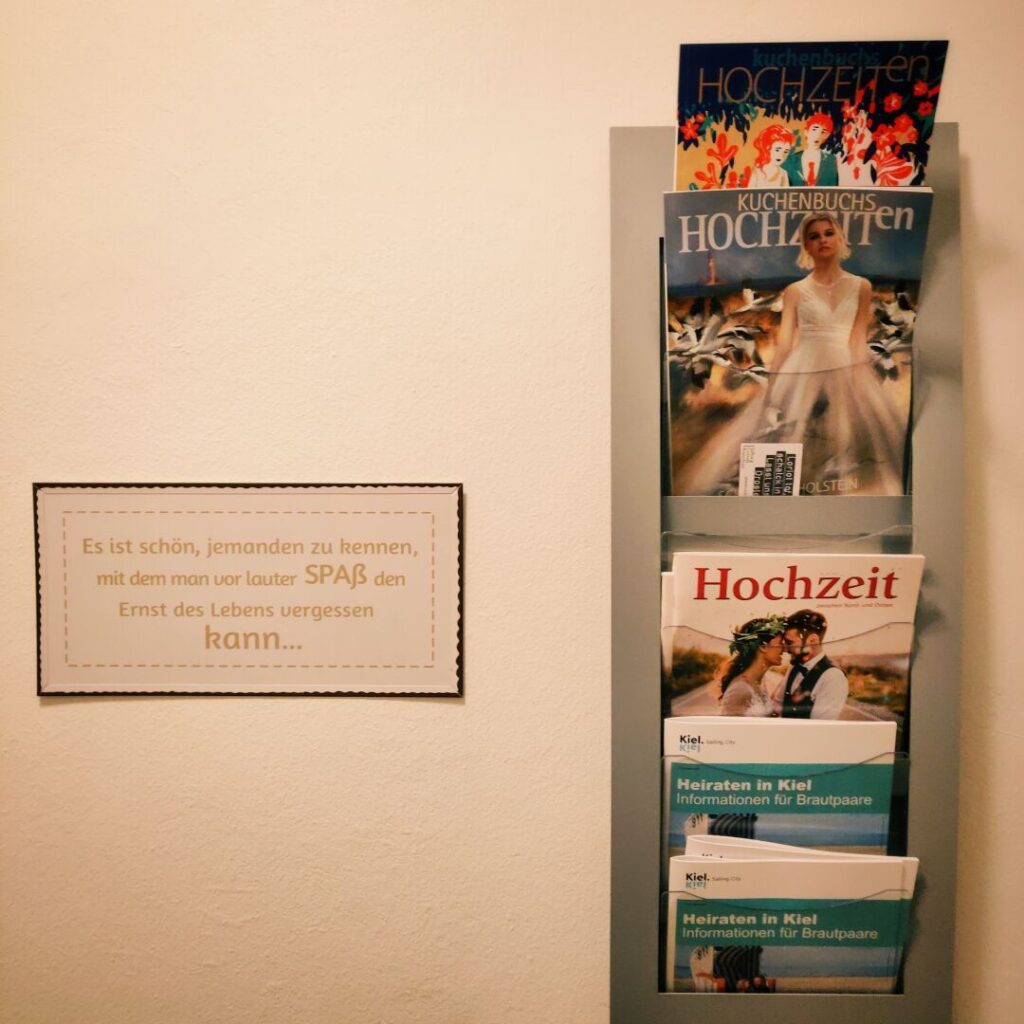 An einer Wand hängt ein Schild mit einem Spruch über Freundschaft und Spaß, daneben befindet sich ein Prospektständer mit Hochzeitsmagazinen und Informationsmaterialien für Brautpaare in Kiel.