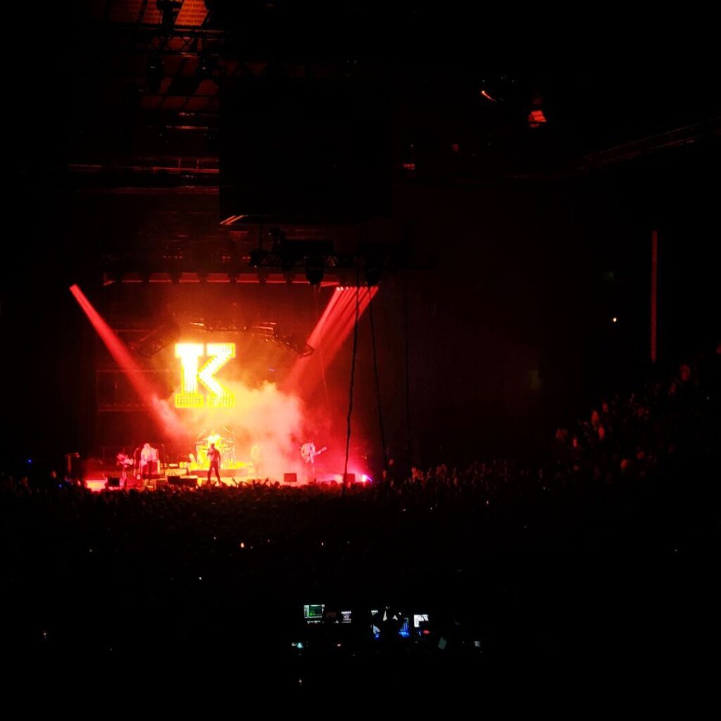 Ein Konzert mit einer großen Menschenmenge, auf der Bühne steht eine Band vor einem leuchtenden Logo, umgeben von bunten Lichtern und Nebel, während die Atmosphäre energiegeladen und festlich wirkt.