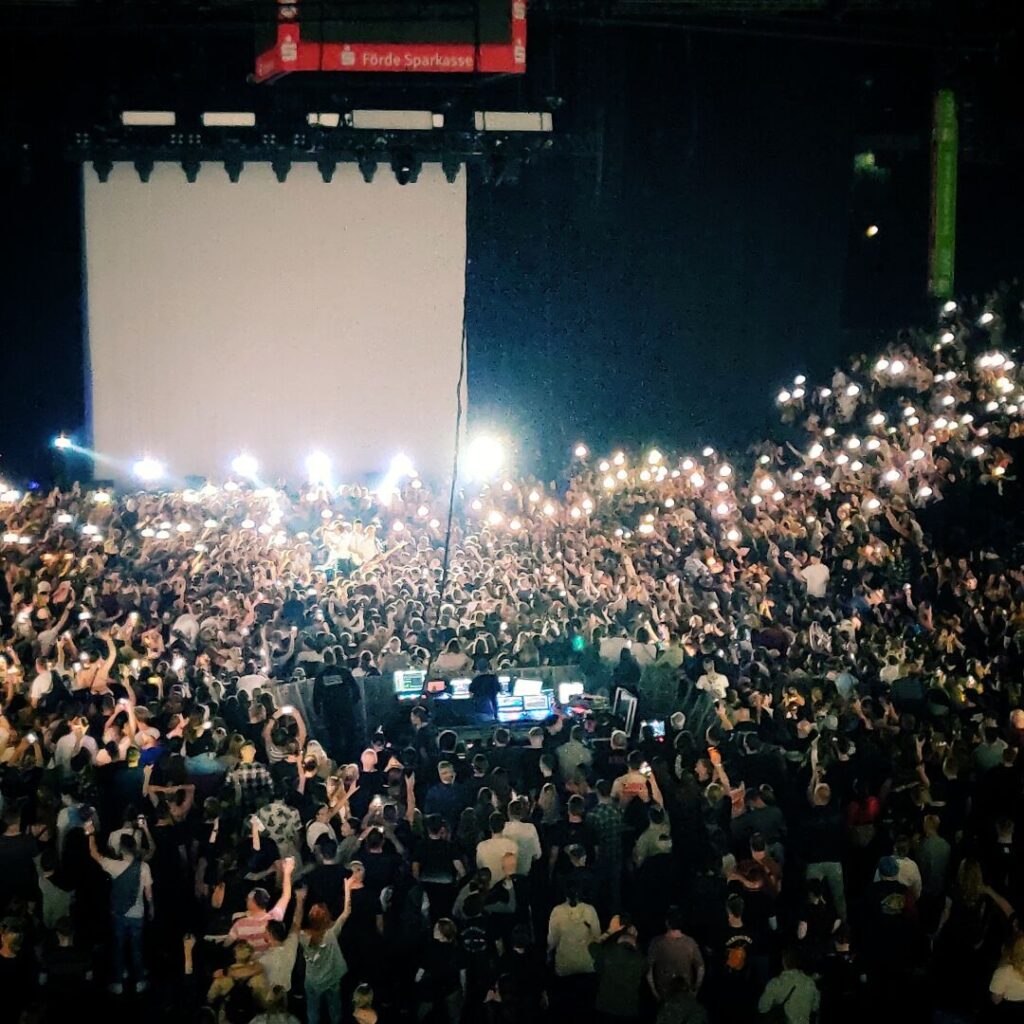 Eine große Menschenmenge mit erhobenen Händen und leuchtenden Handys versammelt sich vor einer Bühne in einem dunklen Veranstaltungsort, während ein Konzert stattfindet.
