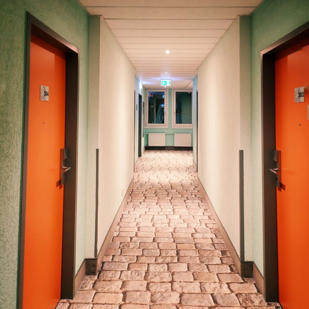 Ein langer, beleuchteter Flur mit orangefarbenen Zimmertüren und einem Fenster am Ende, der mit einem hellen, gepflasterten Boden und grünen Wänden gestaltet ist.
