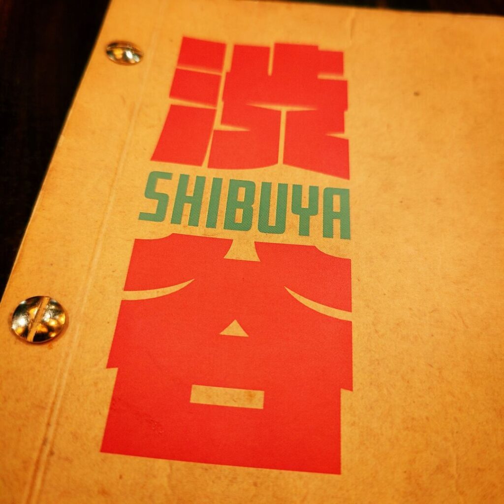 Das Bild zeigt eine Nahaufnahme eines Menüs mit dem Schriftzug "渋谷" in kräftigem Rot und "SHIBUYA" in grün auf einem braunen Hintergrund.