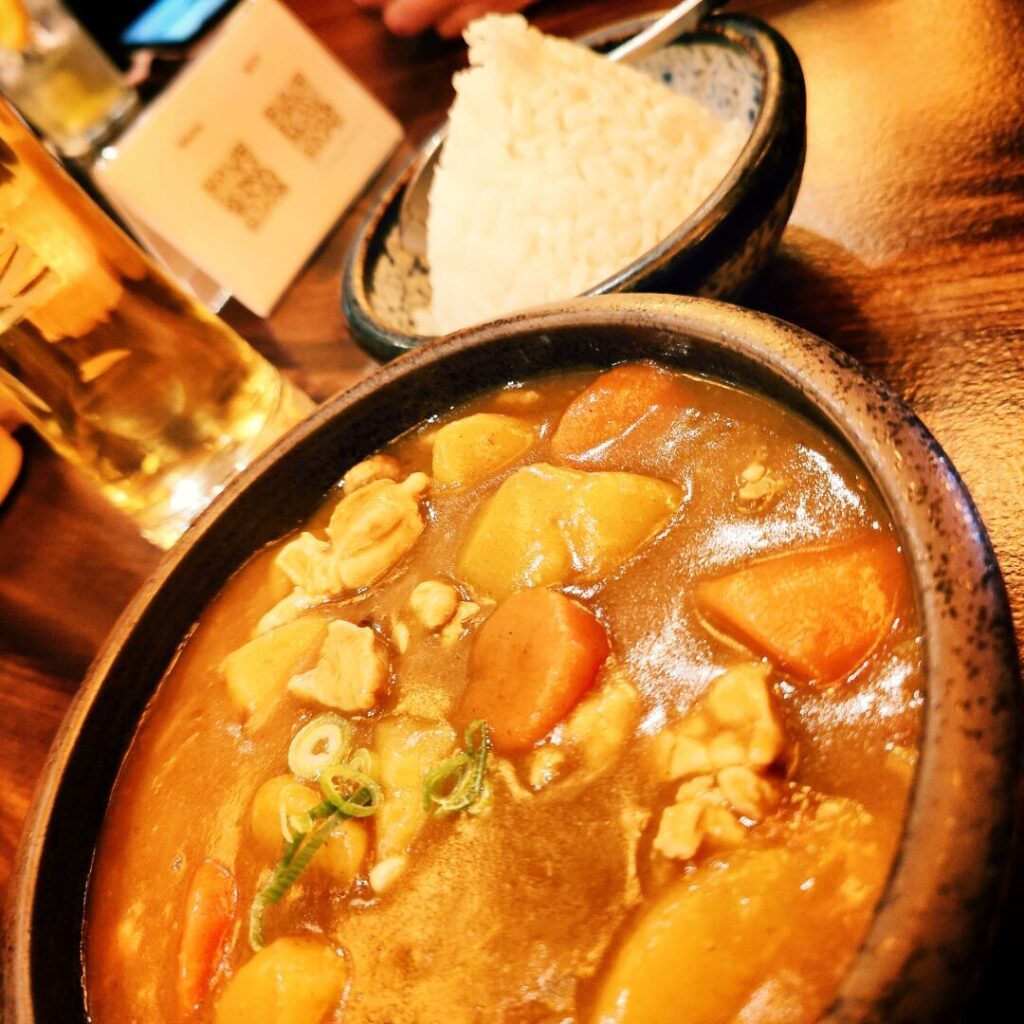Eine Schüssel mit herzhaftem Curry, gefüllt mit Hühnchen, Kartoffeln und Karotten, steht neben einer Portion dampfendem Reis auf einem Holztisch, während im Hintergrund ein Glas Getränk und eine QR-Code-Karte sichtbar sind.