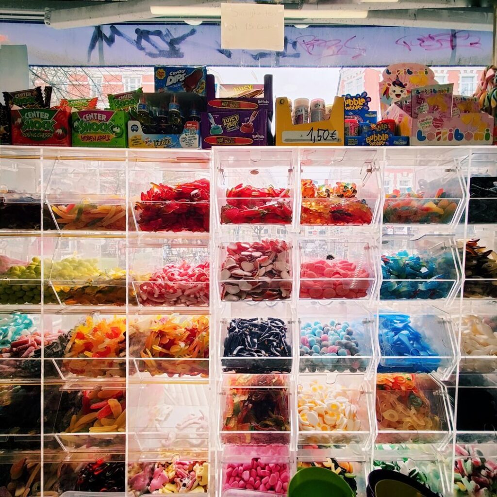 Bunte Süßigkeiten in transparenten Behältern sind in einem Verkaufsstand angeordnet, während im Hintergrund verschiedene Snacks und Getränke präsentiert werden.