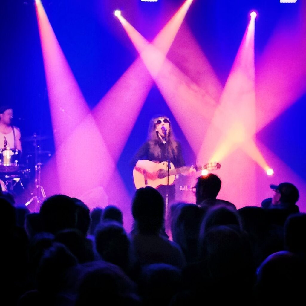 Eine Musikerin mit Sonnenbrille spielt Gitarre und singt auf einer Bühne, umgeben von bunten Lichtstrahlen und einer begeisterten Menge.