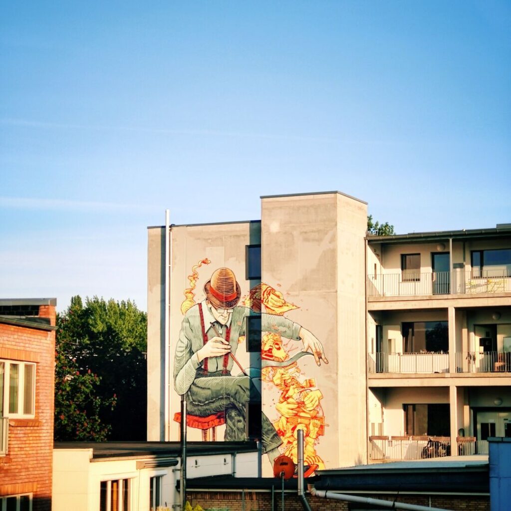 Ein farbenfrohes Wandgemälde zeigt einen älteren Mann mit Hut, der auf einem Stuhl sitzt und mit einer Nadel arbeitet, während um ihn herum lebendige Farben und Formen fließen, vor einem klaren blauen Himmel und modernen Gebäuden.