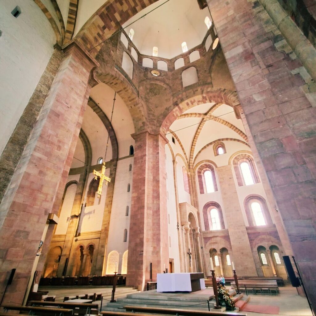 Die Innenansicht einer beeindruckenden Kirche zeigt hohe, mit Steinmauern und Bögen gestaltete Wände, einen zentralen Altar mit einem weißen Tuch und eine goldene Kreuzaufhängung, die das lichtdurchflutete Ambiente betont.