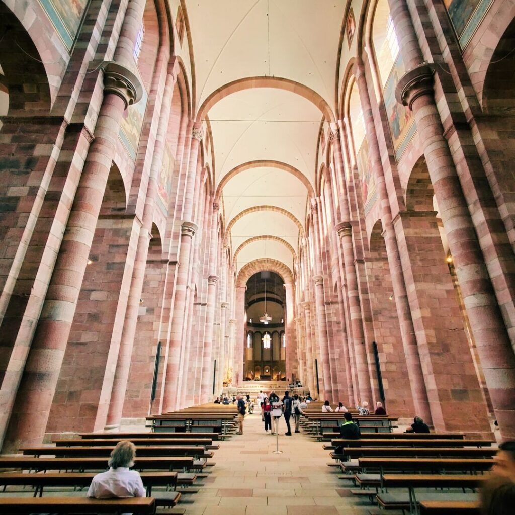 Die Innenansicht einer beeindruckenden Kirche zeigt hohe, gewölbte Decken, massive Säulen aus rosafarbenem Stein und Bänke, auf denen Besucher sitzen und die ruhige Atmosphäre genießen.