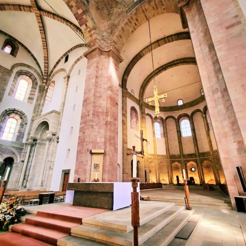 Der Innenraum einer Kirche mit hohen, gewölbten Decken, bunten Fenstern, einem Altarbereich und einer goldenen Kreuzaufhängung, die eine ruhige und spirituelle Atmosphäre vermittelt.