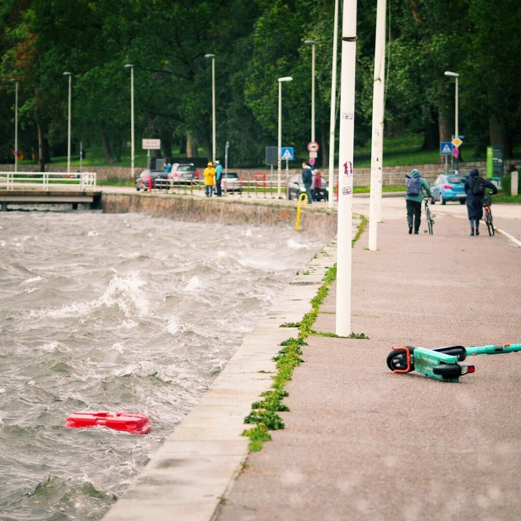 Wellen schlagen gegen die Uferpromenade, während Spaziergänger und Radfahrer bei grauem Wetter vorbeigehen, und ein rotes Objekt schwimmt im Wasser neben einem liegengebliebenen Roller.