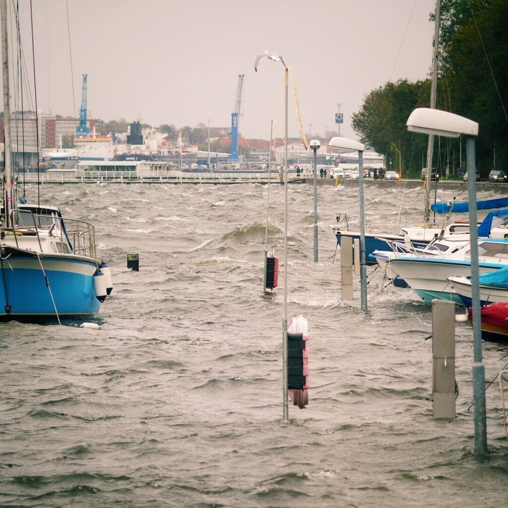 Starkes Hochwasser überflutet einen Hafen mit mehreren Booten, während Wellen gegen die Stege schlagen und im Hintergrund Kräne und Gebäude sichtbar sind.