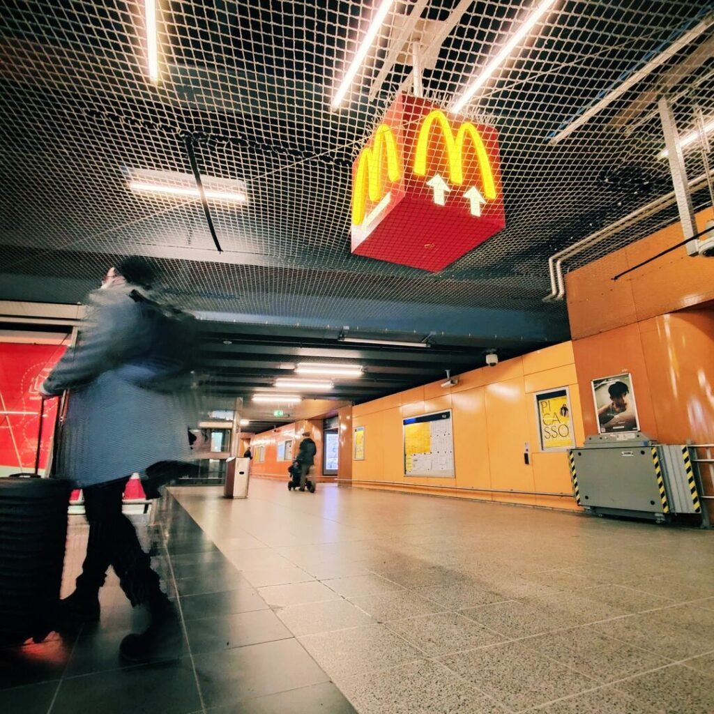 Im Untergeschoss eines Bahnhofs sind Passanten mit Gepäck zu sehen, während ein leuchtendes McDonald's-Schild über ihnen hängt und die orangefarbenen Wände sowie Informationsanzeigen im Hintergrund sichtbar sind.