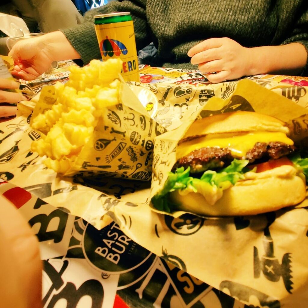 Auf einem bunten Tisch liegen ein saftiger Cheeseburger mit Salat und Tomate sowie eine Portion knuspriger Pommes, daneben steht eine Dose Getränk.