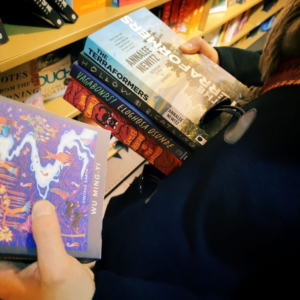 Eine Hand hält mehrere Bücher, darunter "The Terraformers" und "Vagabonds", während ein weiteres Buch mit buntem Cover, "Vintage Earth" von Wu Ming Yi, in der anderen Hand gehalten wird, vor einem Bücherregal voller weiterer Titel.