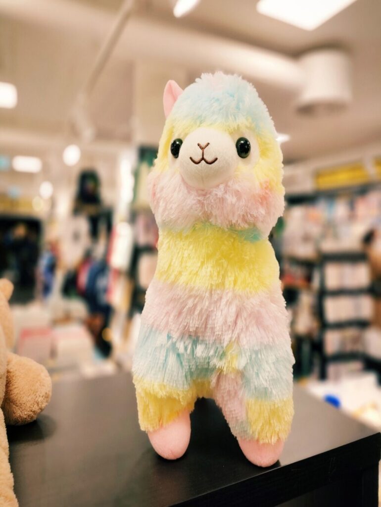 Ein buntes, plüschiges Lama mit pastellfarbenem Fell steht auf einem Regal in einem Geschäft, umgeben von weiteren Spielzeugen und Produkten im Hintergrund.