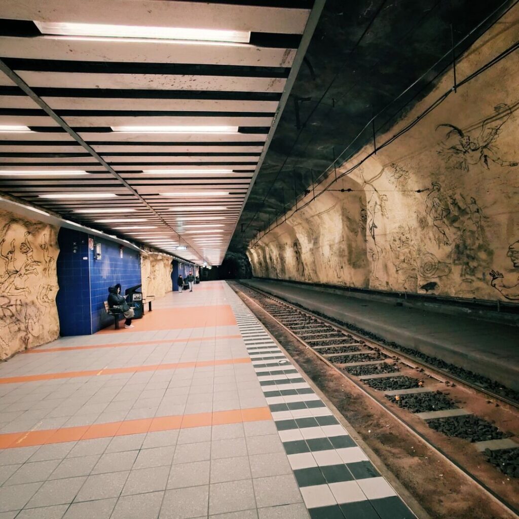 Eine U-Bahn-Station mit langen, leeren Gleisen, einer künstlerisch gestalteten Wand und wenigen wartenden Fahrgästen, die auf einer Bank sitzen, während die Decke mit modernen Lichtleisten beleuchtet wird.