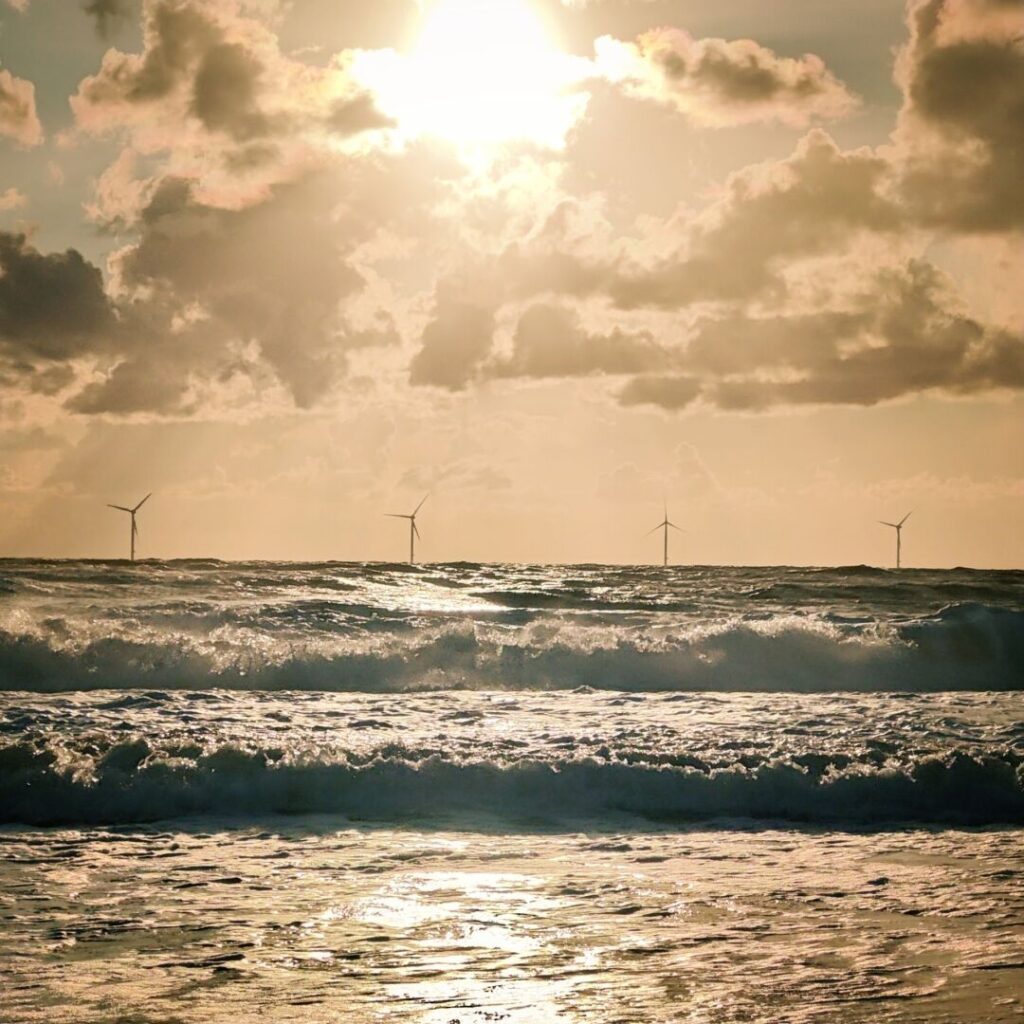Ein Sonnenuntergang über dem Meer mit sanften Wellen und mehreren Windkraftanlagen am Horizont, während die Wolken in warmen Farbtönen leuchten.