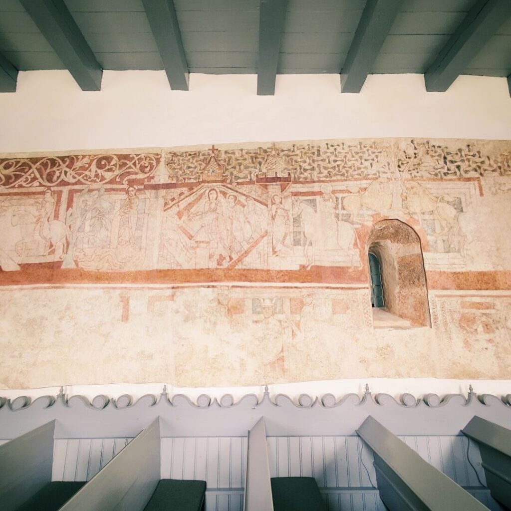 Historische Wandmalereien mit religiösen Motiven und Figuren, die in sanften Farben auf einem hellen Hintergrund dargestellt sind, während im Vordergrund schlichte Sitzbänke zu sehen sind.