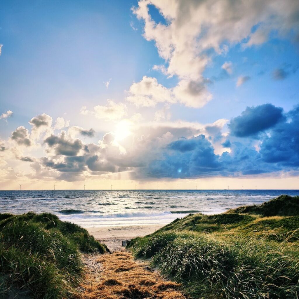 Ein malerischer Blick auf den Strand mit sanften Dünen, grünem Gras und einem strahlenden Sonnenuntergang, während sich die Wellen sanft am Ufer brechen und Windkraftanlagen am Horizont sichtbar sind.