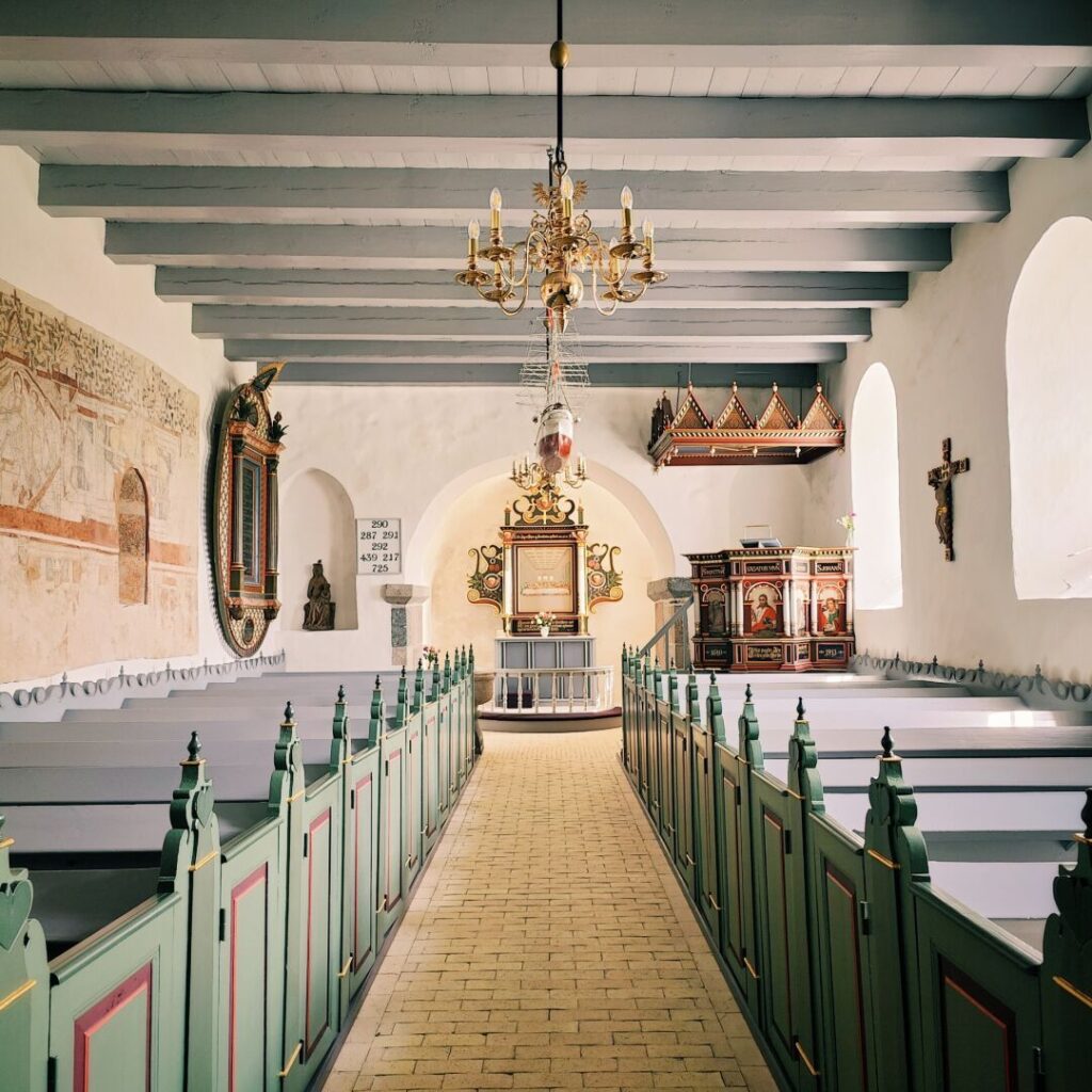Die Innenansicht einer historischen Kirche mit bunten Bänken, einem prächtigen Kronleuchter, kunstvollen Wandmalereien und einem reich verzierten Altar im Hintergrund, der von großen Fenstern umgeben ist.