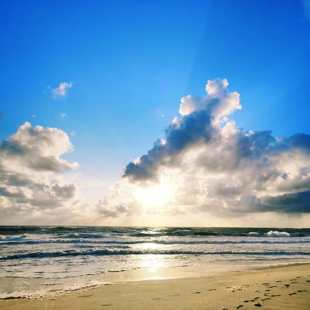 Ein malerischer Strand mit sanften Wellen, die im Sonnenlicht glitzern, während die Sonne hinter einer Wolkenformation am Horizont aufgeht und der Himmel in verschiedenen Blautönen leuchtet.