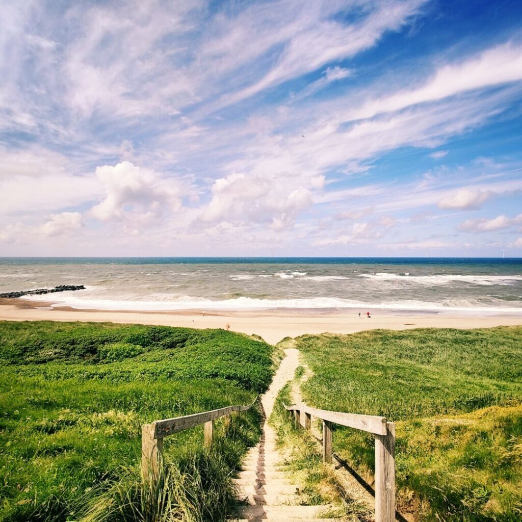 Eine Holztreppe führt durch grünes Gras hinunter zu einem breiten Sandstrand, wo sanfte Wellen auf die Küste treffen, während der Himmel mit weißen Wolken und blauem Hintergrund strahlt.