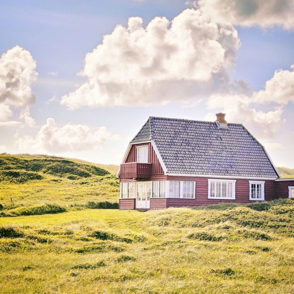Ein rotes Holzhaus mit einem Satteldach steht in einer grünen Wiesenlandschaft unter einem blauen Himmel mit weißen Wolken.
