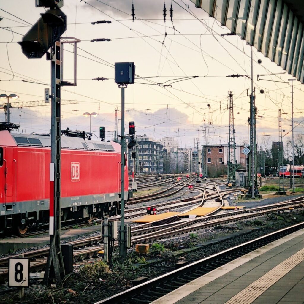 Eine rote Lokomotive der Deutschen Bahn steht an einem Bahnhof, während mehrere Gleise und Signale im Hintergrund zu sehen sind, unter einem bewölkten Himmel mit sanften Lichtverhältnissen.