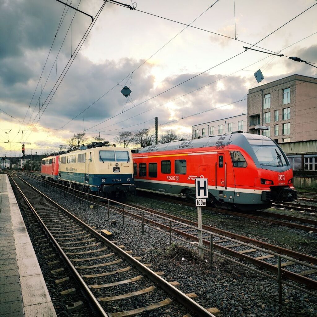 Zwei Züge stehen auf einem Bahnhofsgleis, ein älterer, blau-weißer Lokomotive und ein moderner, roter Triebwagen, während sich im Hintergrund ein bewölkter Himmel über einem Gebäude erstreckt.