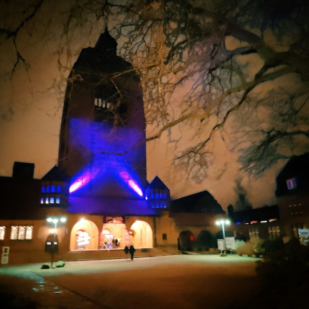 Die beleuchtete Fassade eines historischen Gebäudes mit einem hohen Turm, umgeben von Bäumen, während die Nacht hereinbricht und die Atmosphäre durch sanfte Lichter in Blau und Violett verstärkt wird.