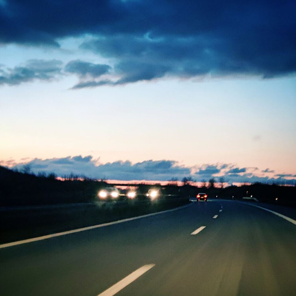 Eine Autobahn mit mehreren Fahrzeugen bei Dämmerung, umgeben von einem farbenfrohen Himmel mit Wolken und einem sanften Lichtspiel am Horizont.