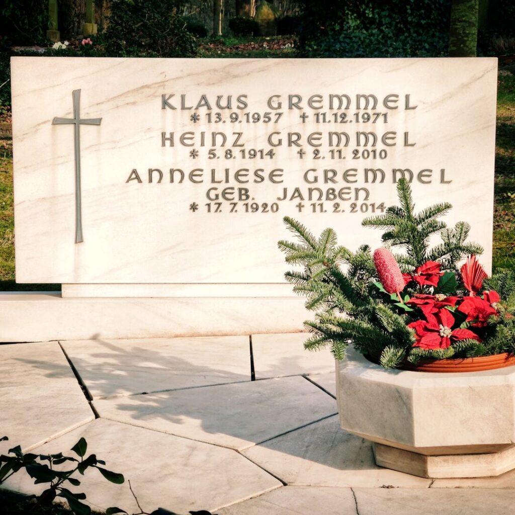 Das Grabmal der Familie Gremmel zeigt die Namen Klaus, Heinz und Anneliese Gremmel sowie ihre Geburts- und Sterbedaten, umgeben von einer dekorativen Pflanzschale mit roten Blüten und grünen Zweigen.