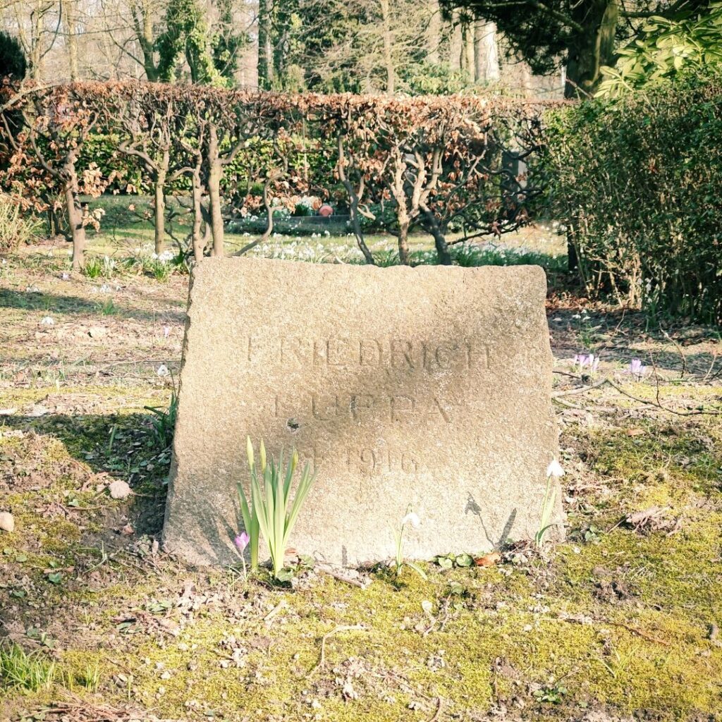 Ein gravierter Grabstein mit der Inschrift "Friedrich Lueppa 1910" steht in einem ruhigen Garten, umgeben von frischen Pflanzen und blühenden Krokussen, während die Sonne durch die Bäume scheint.