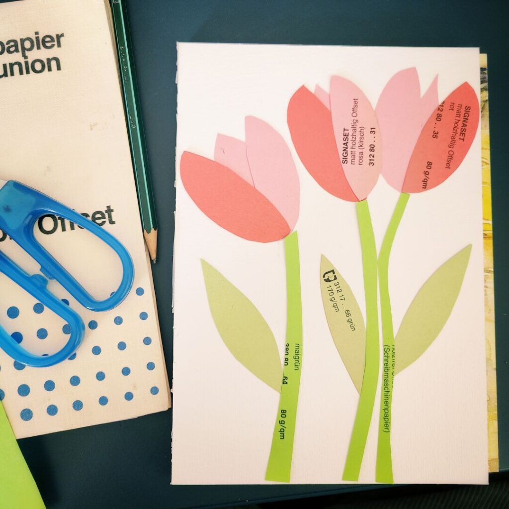 Bunte Papierblumen aus verschiedenen Materialien auf einem weißen Hintergrund, umgeben von Bastelutensilien wie einer Schere, einem Bleistift und einem Buch über Offsetpapier.