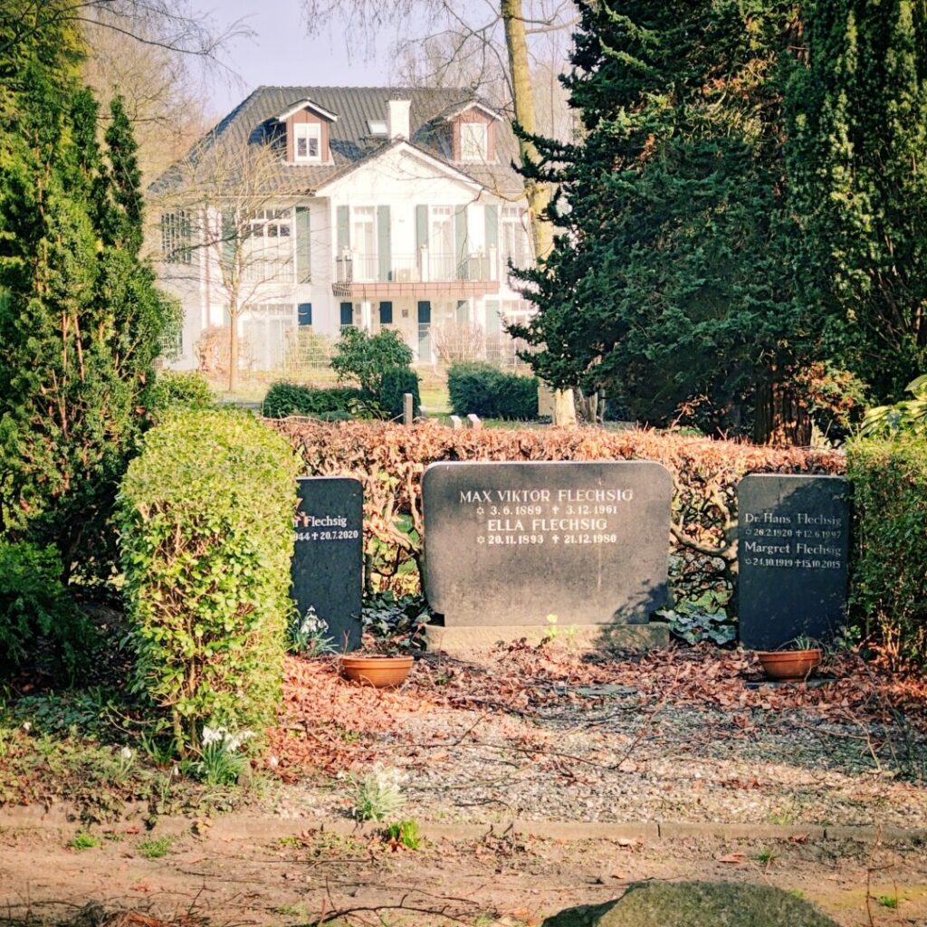 Gräber von Max Viktor Flechsig und Ella Flechsig in einem ruhigen Garten mit einem modernen Haus im Hintergrund, umgeben von grünen Sträuchern und frischen Pflanzen.